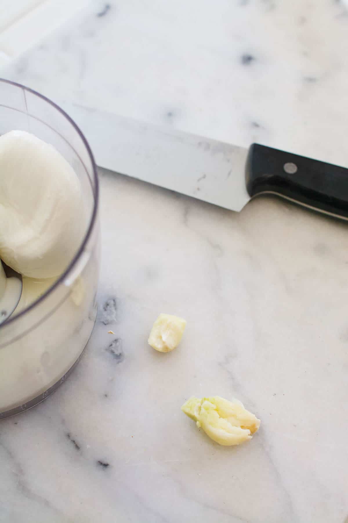 A smashed clove of garlic on a marble counter top between a kitchen knife and a food processor bowl.