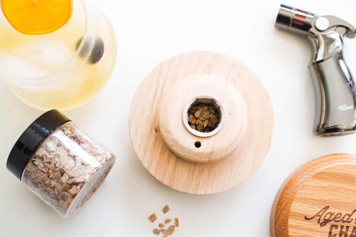 A cocktail smoker kit from Aged and Charred including a cocktail smoker, wood chips and a butane torch next to a highball glass.