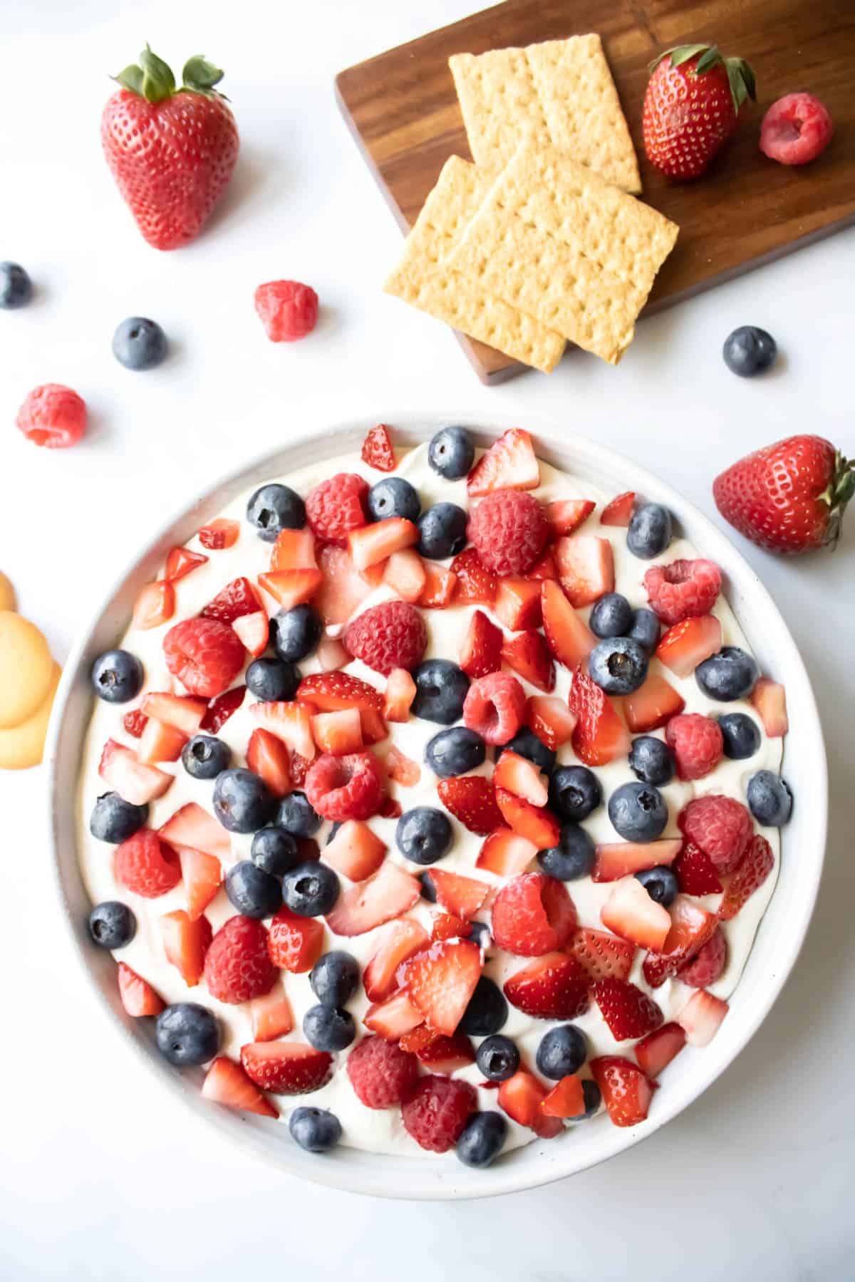 Cheesecake fruit dip in a bowl topped with red and blue berries and dippers like graham crackers and cookies.