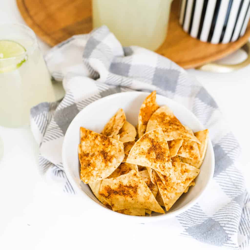 Close up image of spiced tortilla chips in a white bowl on top of a gray checked towel.