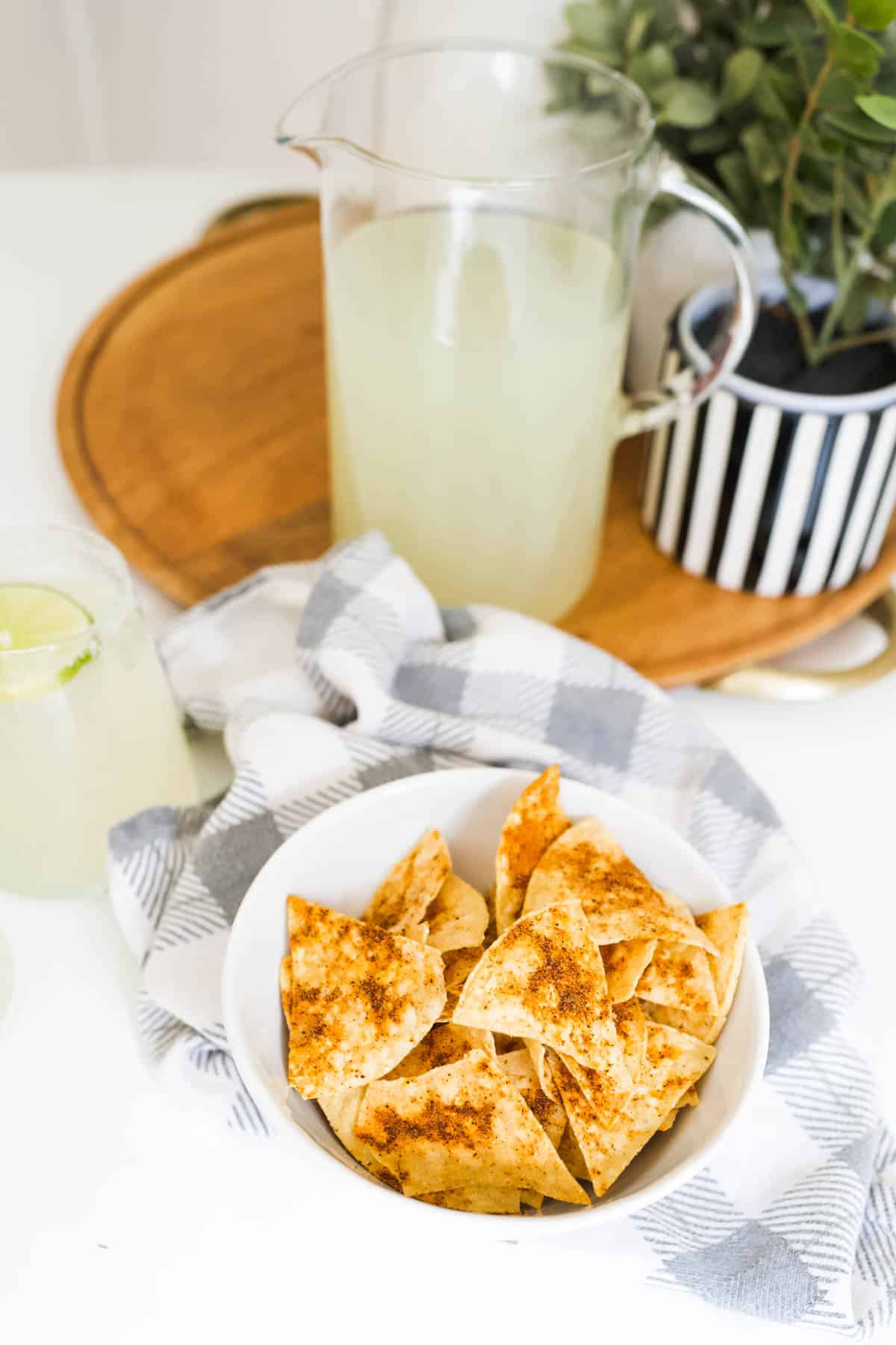 A white bowl filled with spiced tortilla chips on top of a kitchen towel next to a pitcher of lemonade on a wooden tray.
