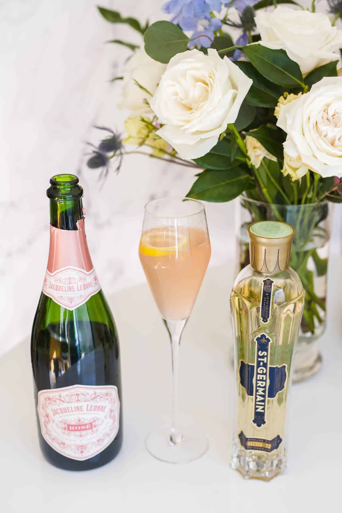 Bottles of pink champagne and St Germain next to a champagne cocktail with a vase of flowers in the background.