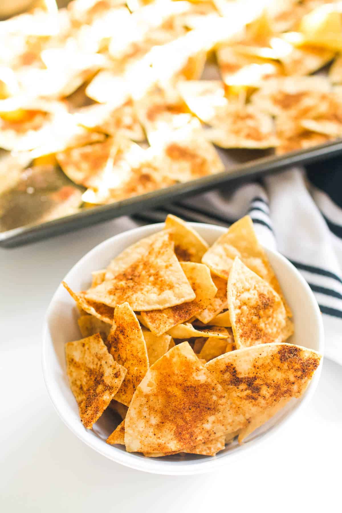 Store bought tortilla chips seasoned with spice mix in a white bowl, next to a sheet pan filled with tortilla chips.