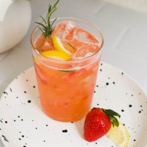 A strawberry and rosemary mocktail in a glass topped with fresh rosemary and lemon next to fresh fruit on a plate.