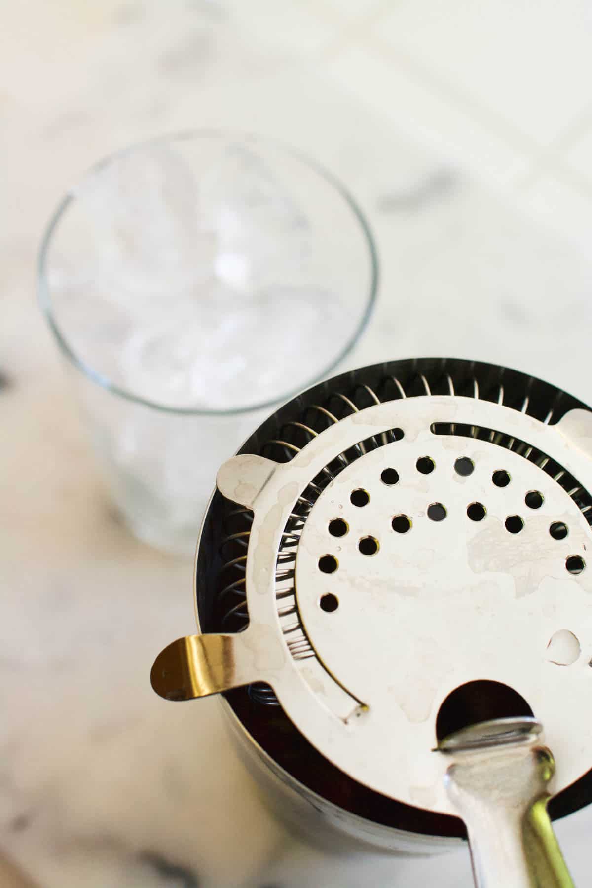 A glass of ice next to a cocktail shaker topped with a strainer.