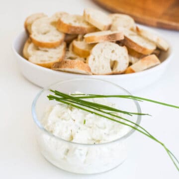 A small glass bowl of truffle burrata dip topped with chives in front of a bowl filled with crostini.