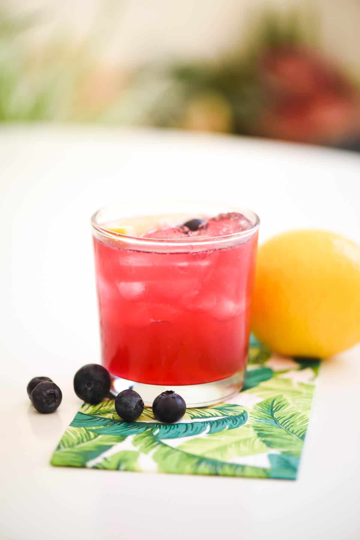 A blueberry lemonade cocktail with vodka in a high ball glass and surrounded by fresh lemon and blueberries.