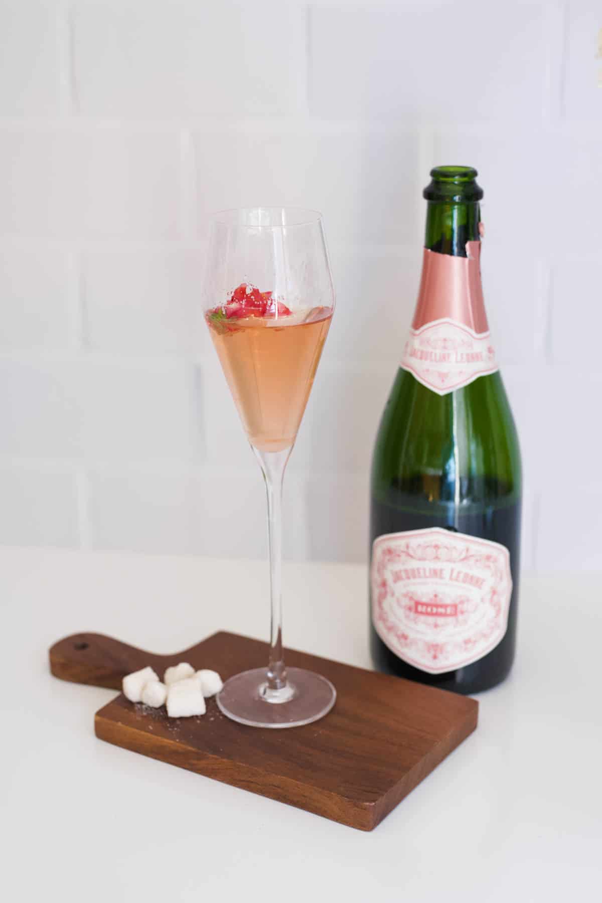 A champagne cocktail on a wooden board with sugar cubes next to a half empty bottle of pink champagne.