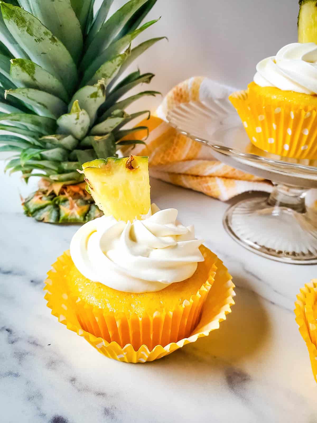 A single pineapple cupcake topped with fresh pineapple in front of a cake stand holding another cupcake and a pineapple top.