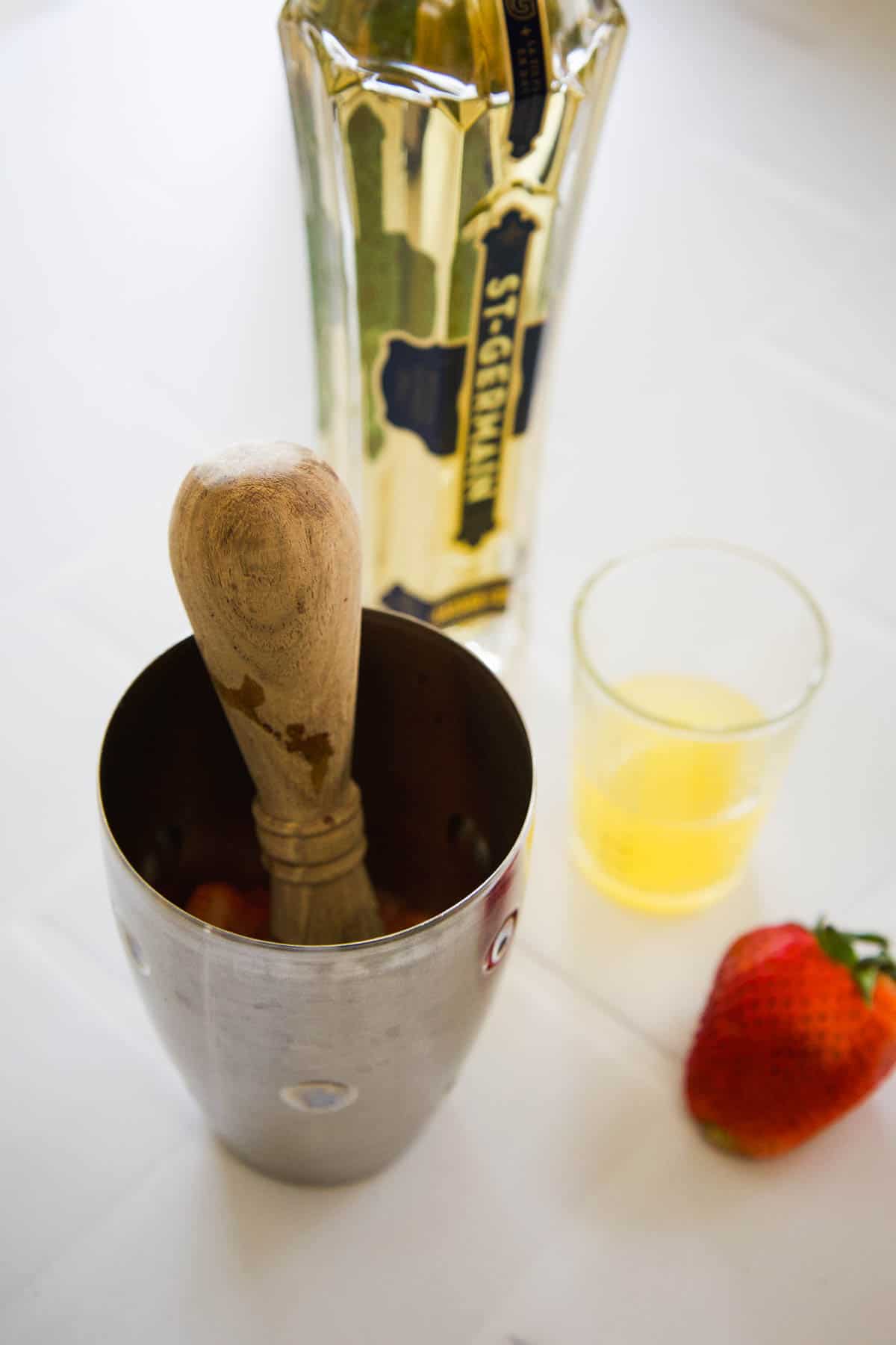 A cocktail shaker with a muddler in it next to a St Germain bottle, a measuring cup with lemon juice and a strawberry.