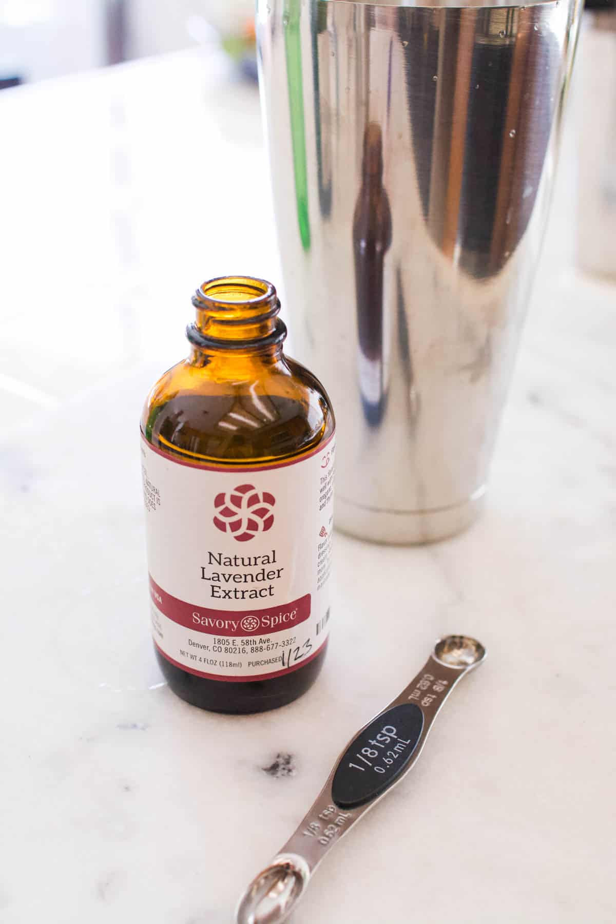 Bottle of lavender extract next to a cocktail shaker with a measuring spoon.