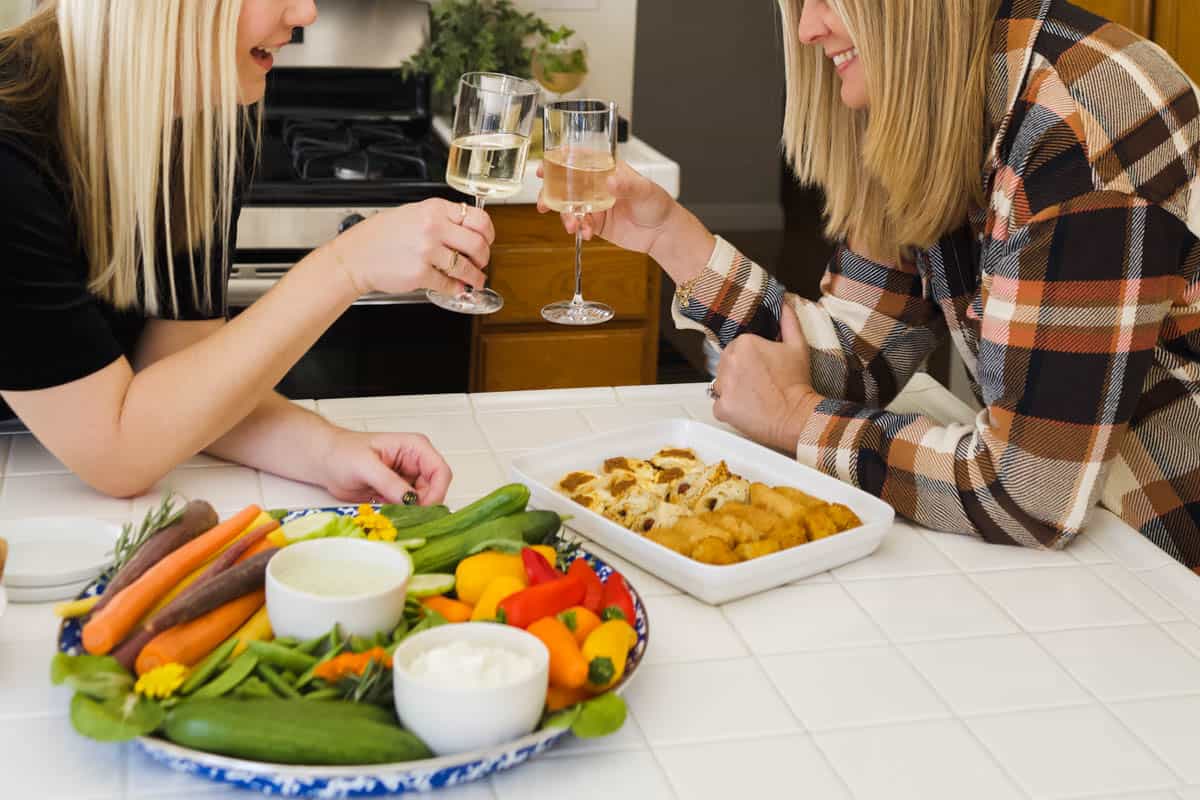 Two woman cheersing wine glasses with a try of appetizers and dips on the counter in front of them.