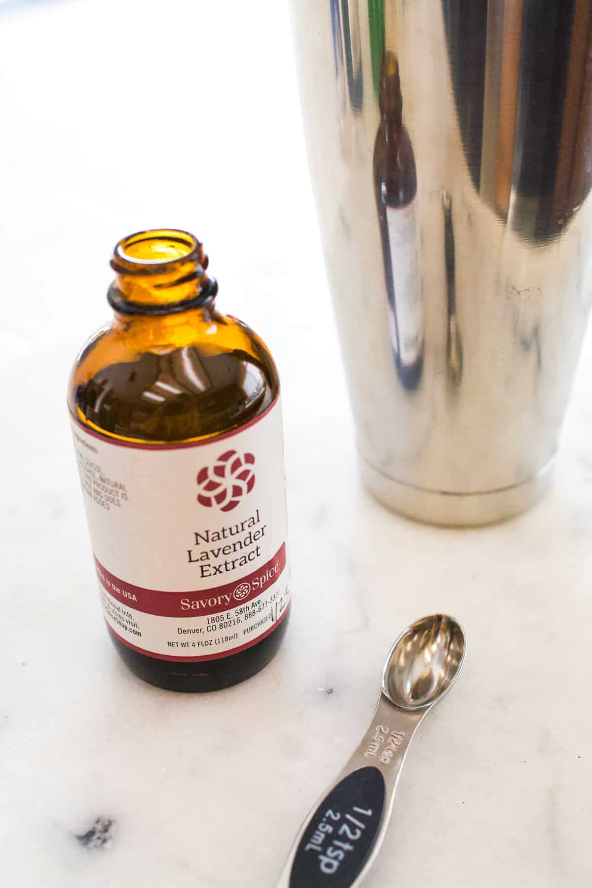 Bottle of lavender extract and a measuring spoon next to a cocktail shaker.