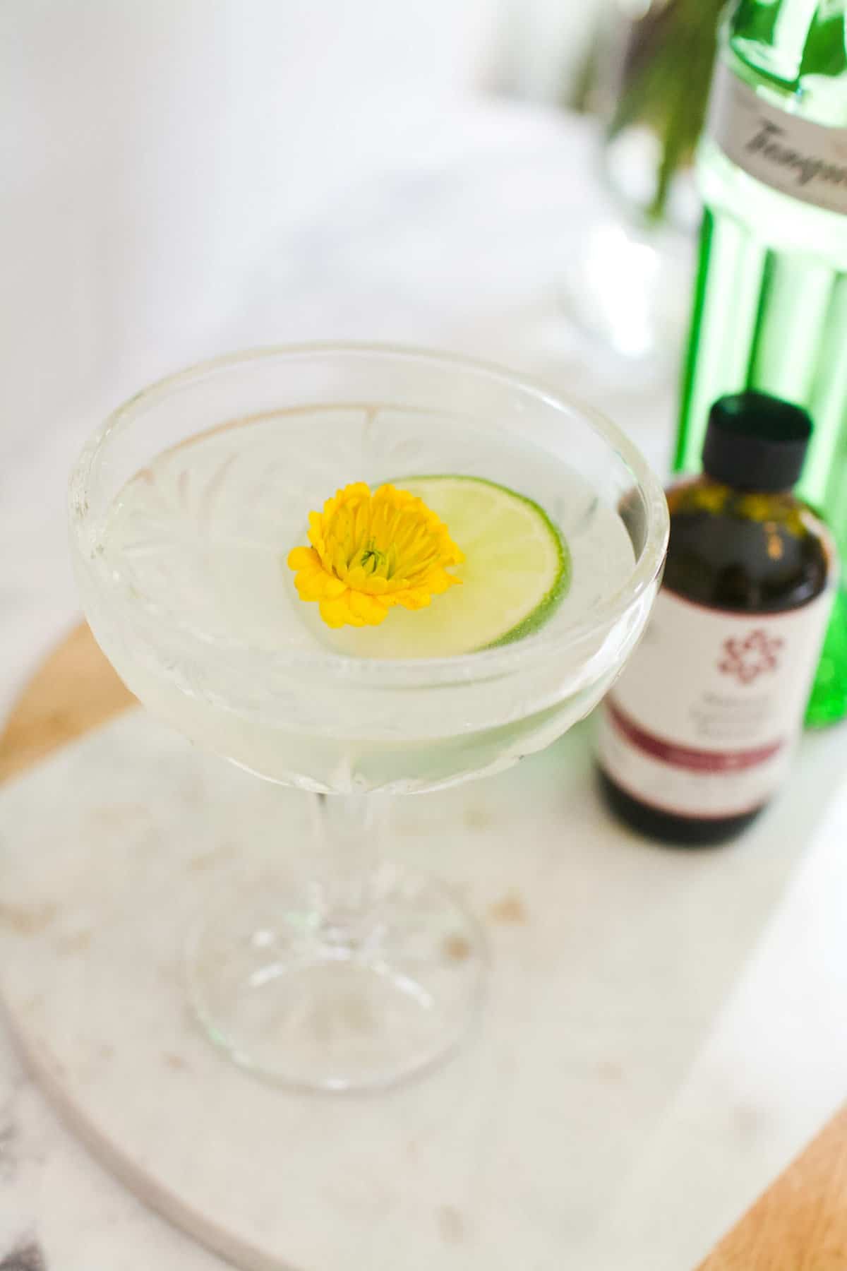 A gimlet garnished with a lime wheel and edible flower with a bottle of lavender bitters and gin in the background.