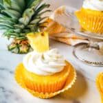 A pineapple cupcake on a white counter top next to the top of a pineapple and a cake stand with more cupcakes.