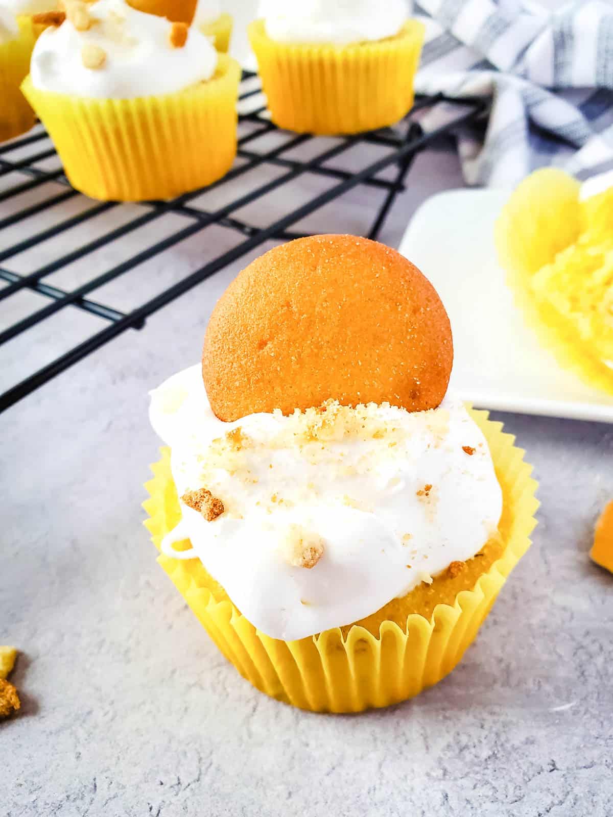 A Banana Pudding cupcake in front of a cooling rack holding more cupcakes.