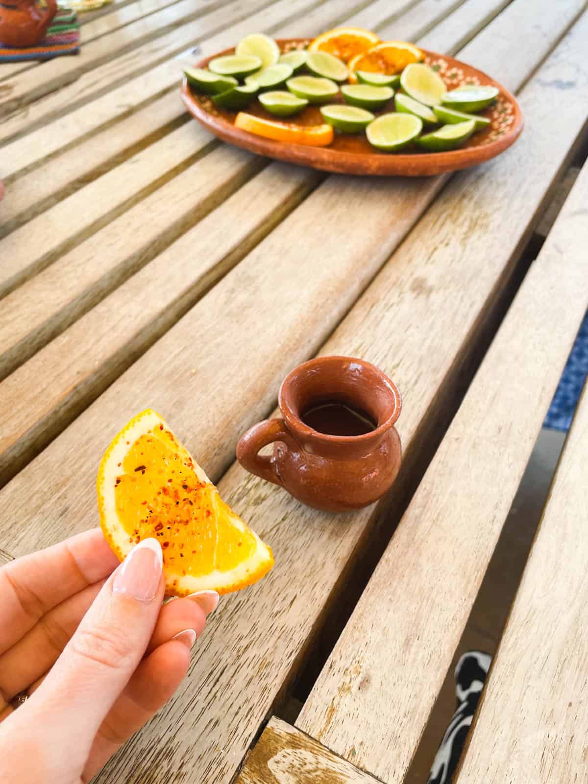 Woman holding a slice of orange with chile on it next to a shot of tequila in a terra cotta shot glass.