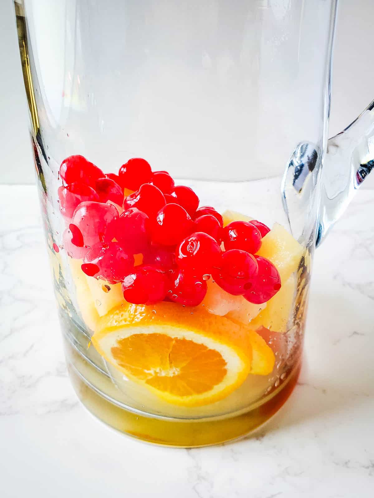 A pitcher with sliced oranges, chunks of pineapple and marashino cherries.