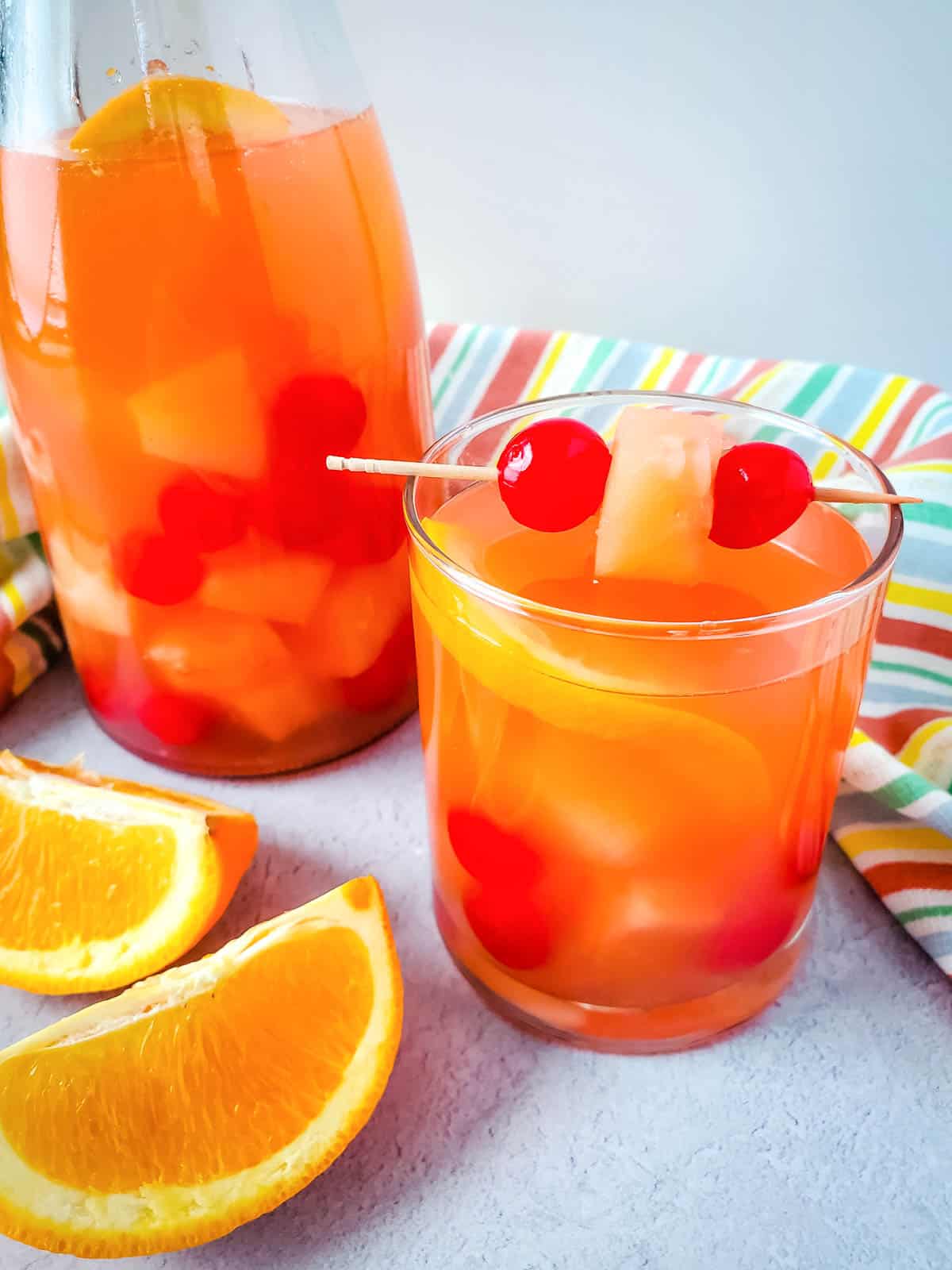 A glass of passion fruit sangria with spiced rum with garnish of cherries and pineapple chunk next to a pitcher of sanria.