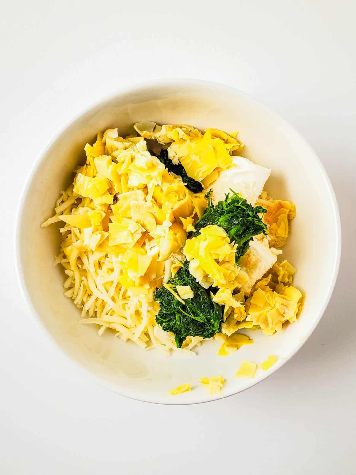 A white bowl with ingredients to make a quick spinach artichoke dip to top on a bread.