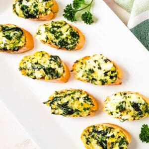 Close up of a white platter with baked crostini topped with spinach artichoke dip.