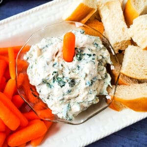 Spinach dip with a carrot on top on a white serving plate between carrots and bread.