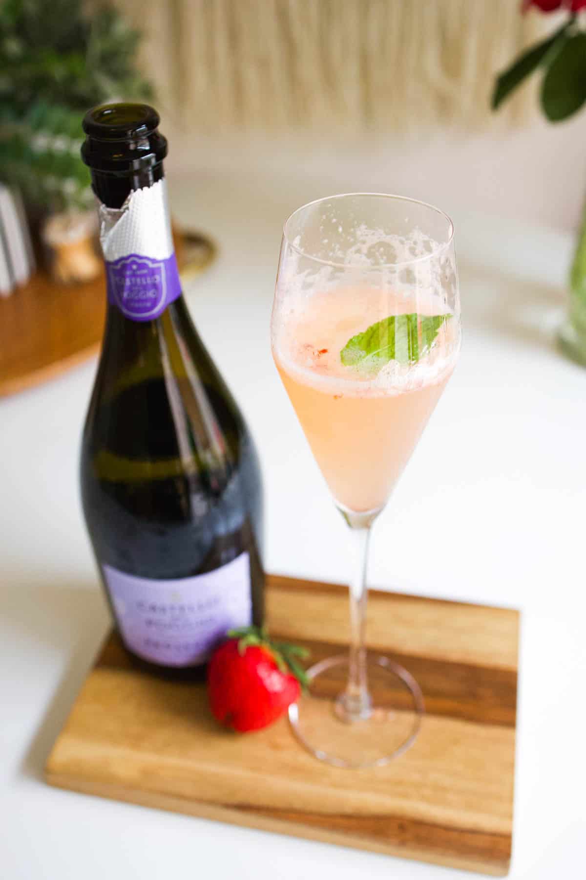 A champagne flute on a wooden board with a cocktail in it next to a strawberry and bottle of prosecco.