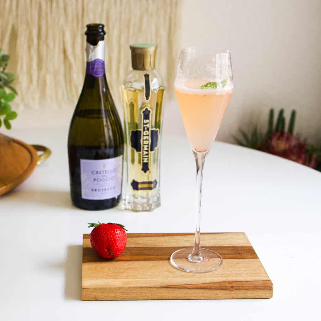 A wooden board on a table holding a strawberry Prosecco cocktail with a bottle of prosecco and st germain behind it.