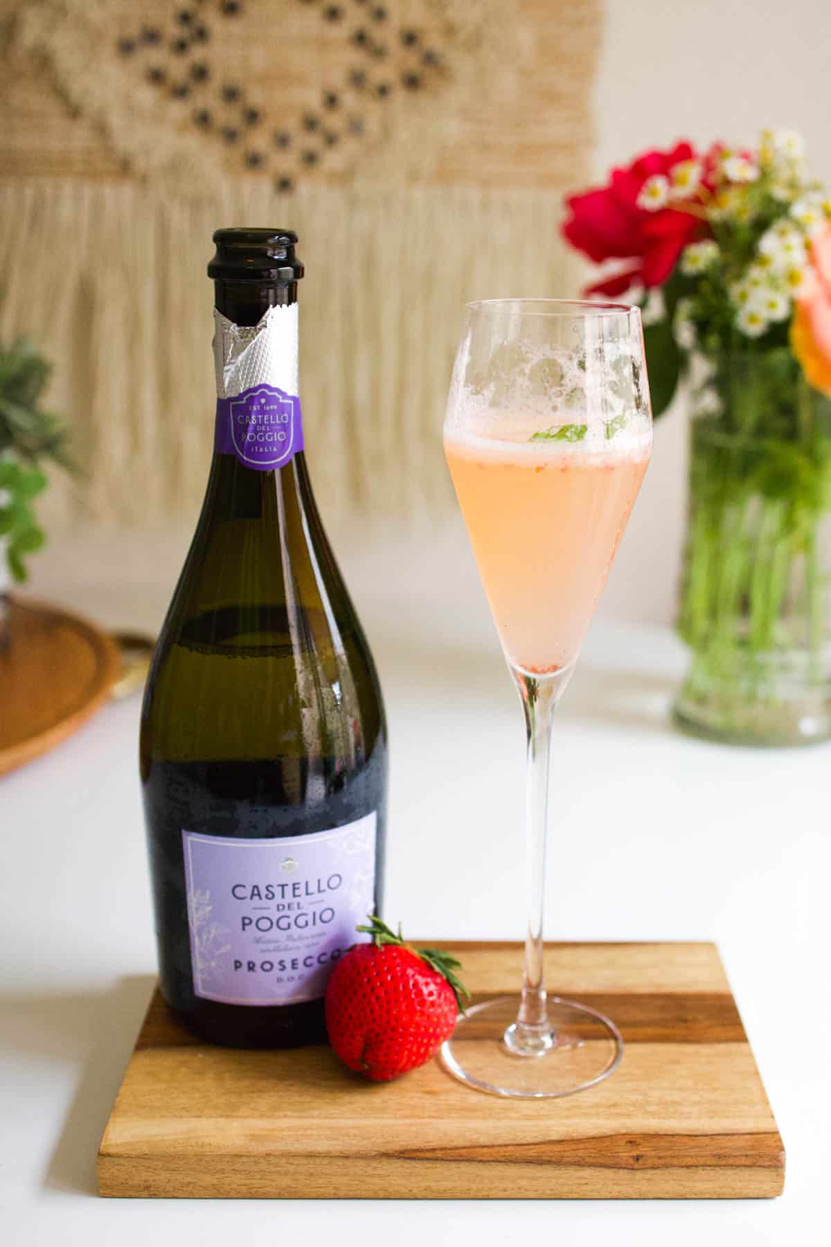 A bottle of prosecco on a wooden tray with a champagne flute with a cocktail and a fresh strawberry.