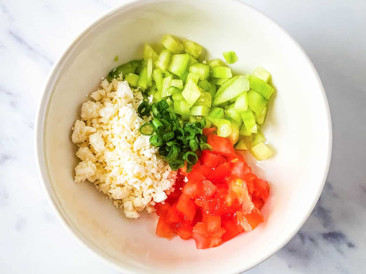 A bowl with chopped cucumbers and tomatoes with feta and green onion.