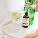 A gin gimlet using lavender extract served in a crystal coupe glass on a table next to gin and a bottle of lavender extract.