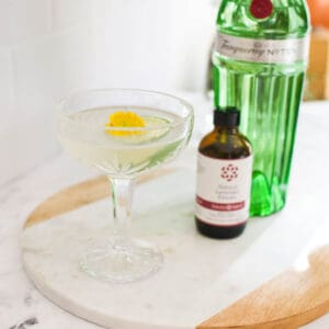 A gin gimlet using lavender extract served in a crystal coupe glass on a table next to gin and a bottle of lavender extract.