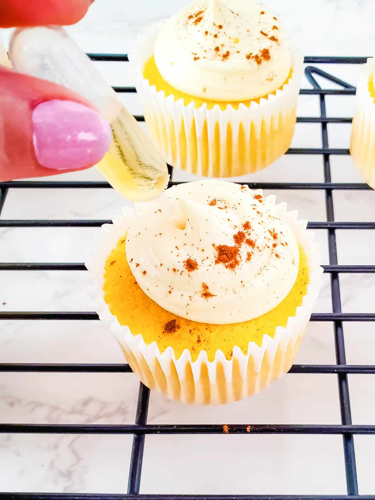 Fingers adding a pipette with spiced rum into cupcakes with frosting and cinnamon on a wire rack.