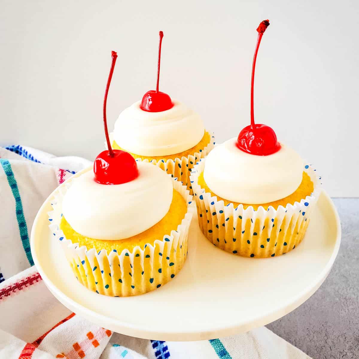 Three coconut cupcakes with frosting and maraschino cherries on a platter.