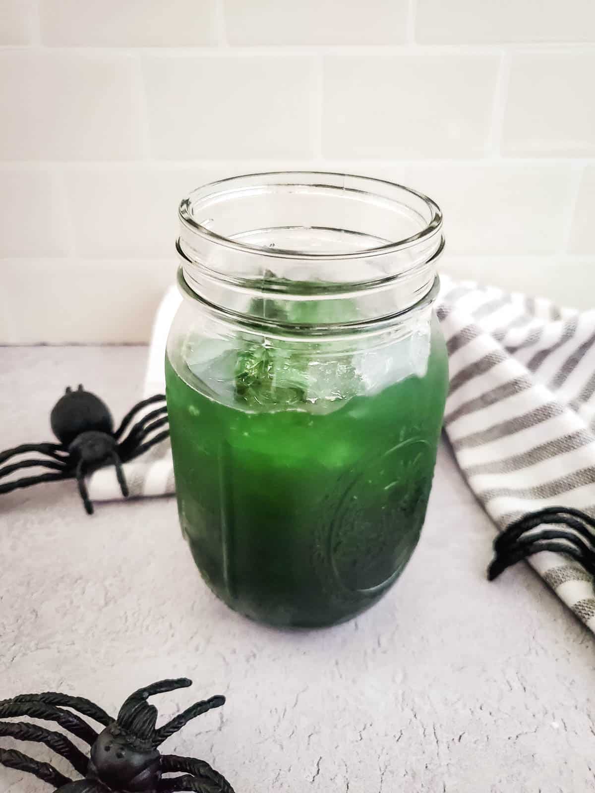 Green halloween cocktail in a mason jar surrounded by plastic spiders.