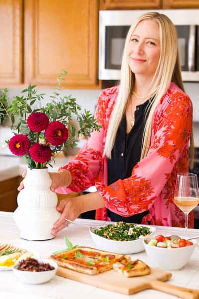 Woman setting a vase of flowers on a counter with food and wine on it.