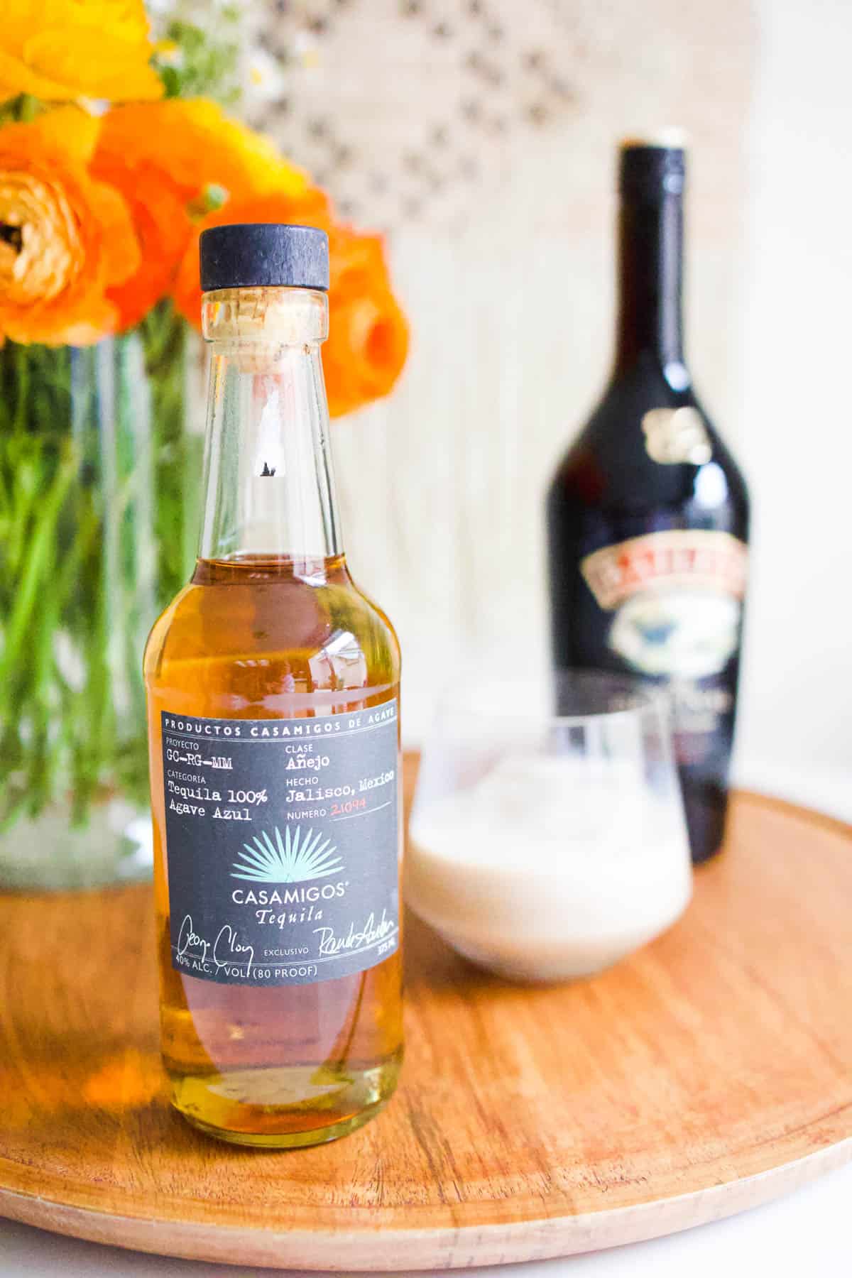 A bottle of reposado tequila on a tray with a bottle of Baileys in the background and a cocktail glass on a tray.