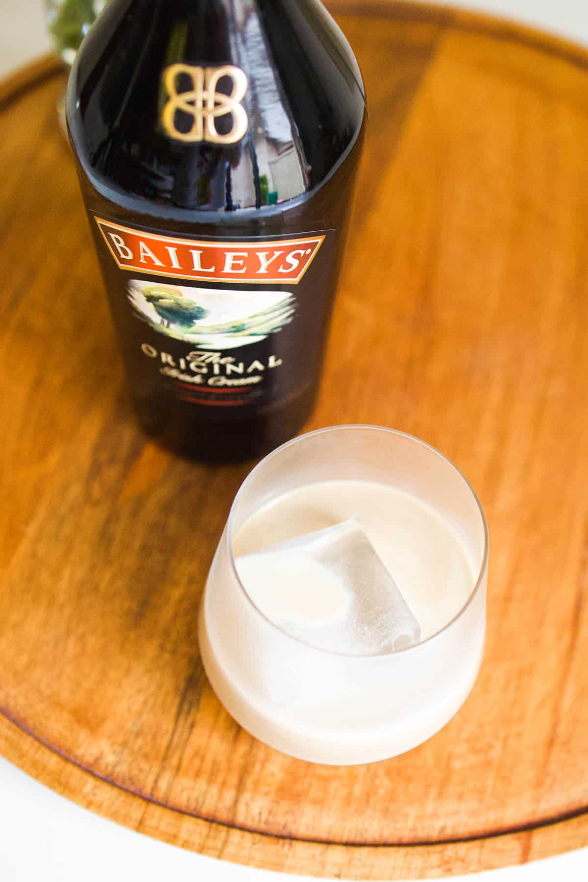 A glass on a wooden tray with a large ice cube and Baileys Irish Cream next to a bottle of Baileys.