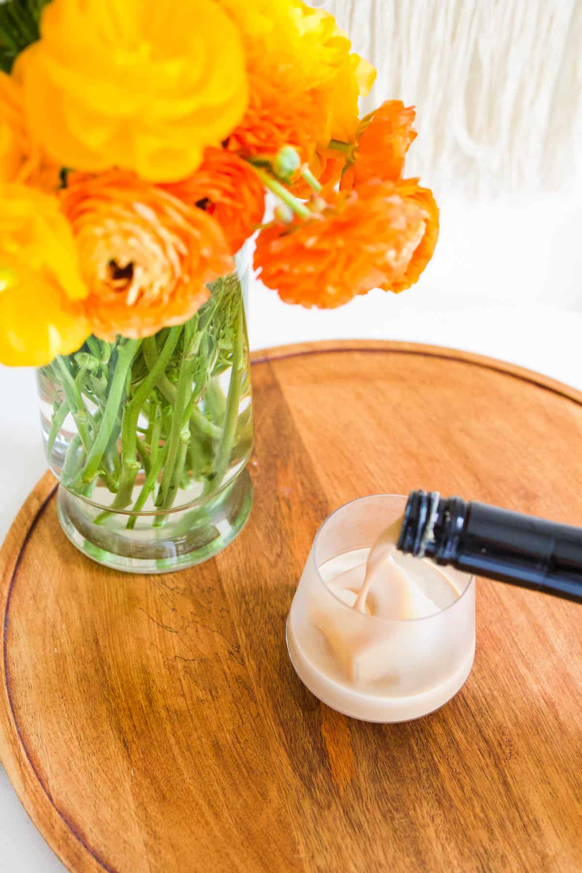 A vases of flowers on a wooden tray with someone pouring Baileys out of the bottle over a large ice cube in a glass.