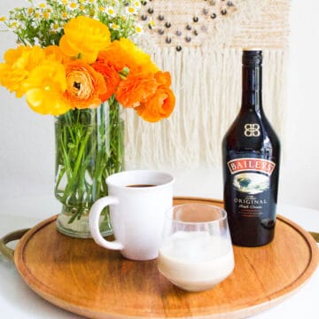 A wooden tray with a bottle of Baileys next to a cup of coffee and a glass with Baileys and a large ice cube and a vase of flowers.