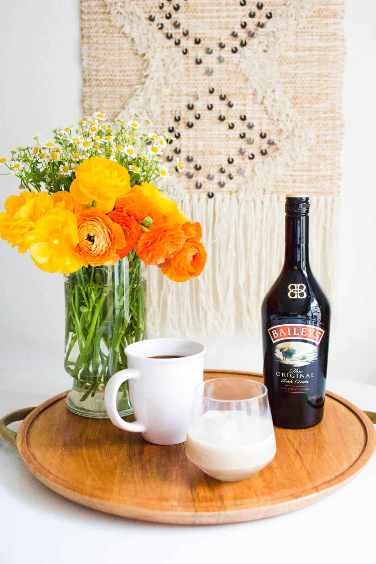 A tray on a table with a vase of flowers and a bottle of Irish Cream next to a cup of coffee and a cocktail glass.