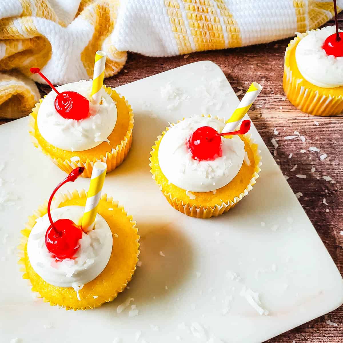 Boozy pina colada cupcakes with vanilla frosting and maraschino cherries on a plate.