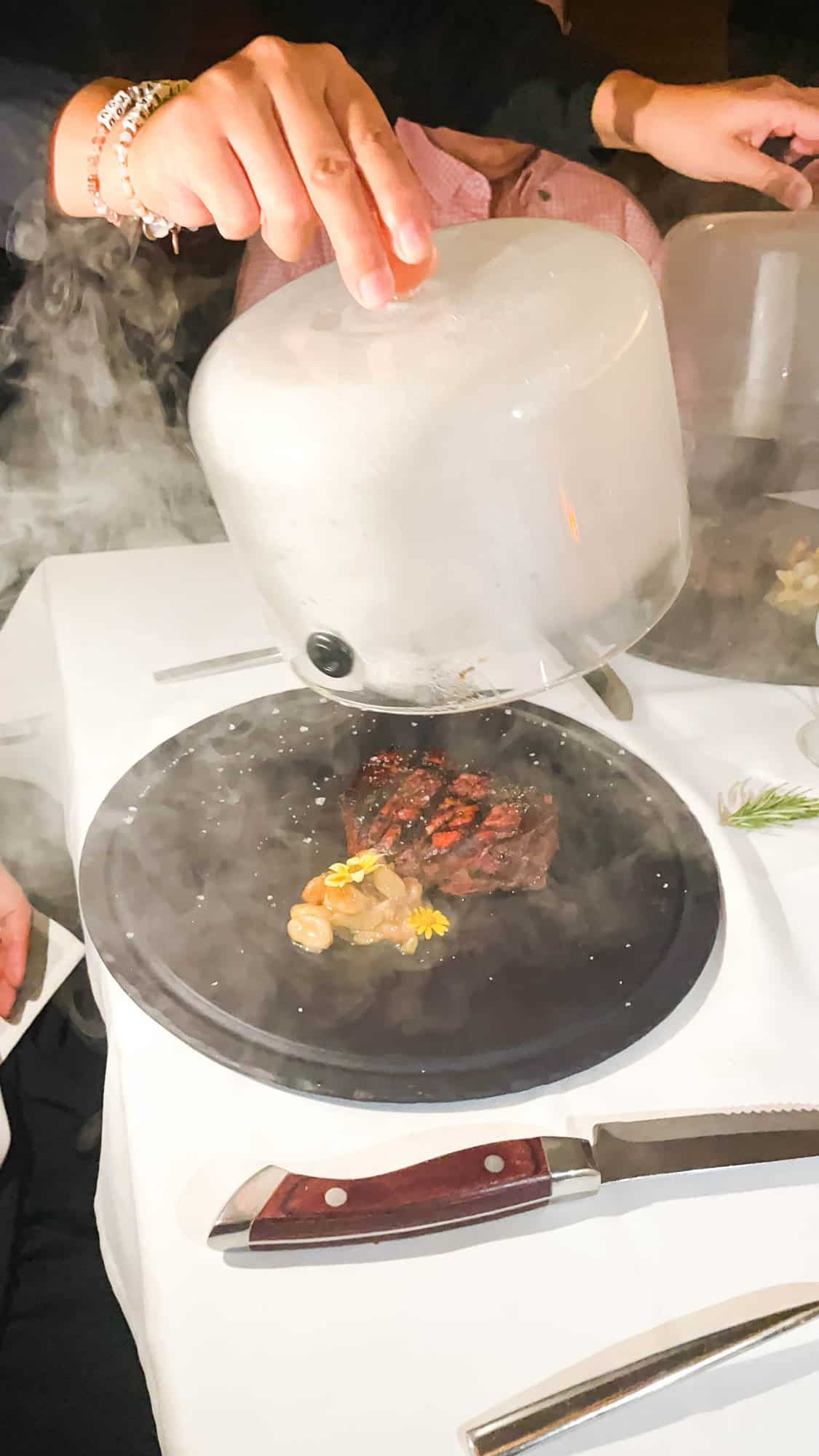 A dinner plate with a steak with a lid being lifted off of the plate and smoke coming all around it.