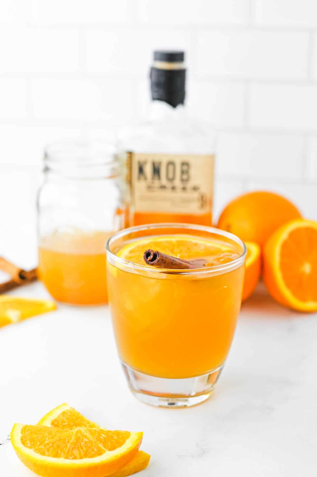 An apple cider old fashioned surrounded by oranges, a jar of apple cider and a bottle of bourbon on a white countertop.