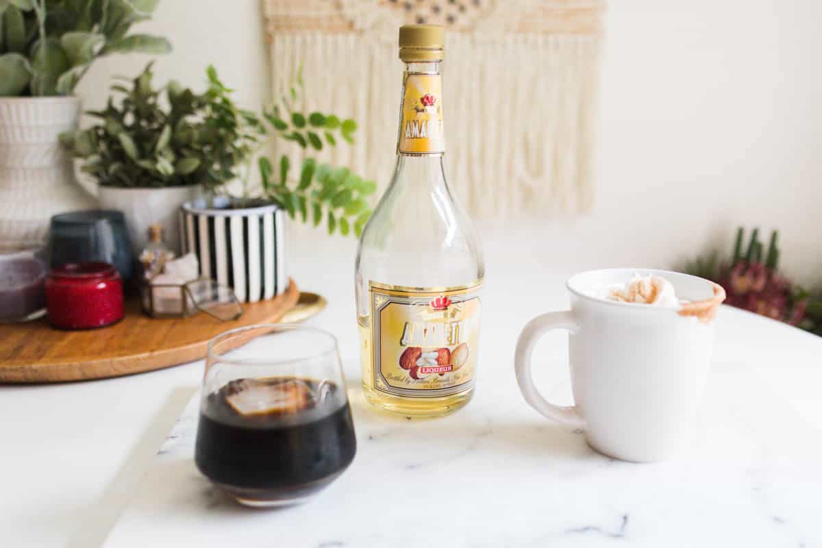An iced coffee and a mug of coffee next to a bottle of amaretto.