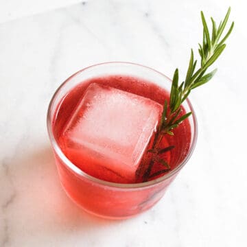 Cranberry and tequila cocktail over a giant ice cube garnished with a fresh rosemary sprig.