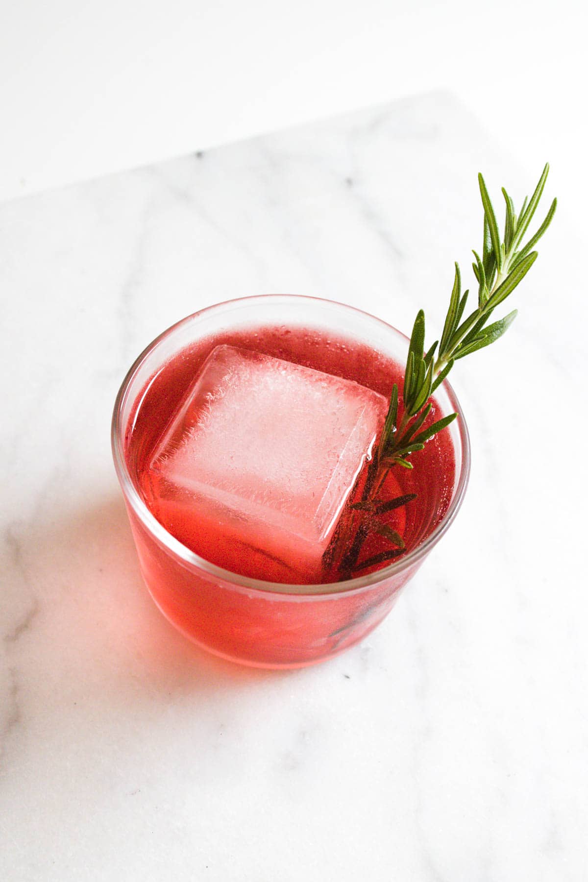 Cranberry and tequila cocktail over a giant ice cube garnished with a fresh rosemary sprig.