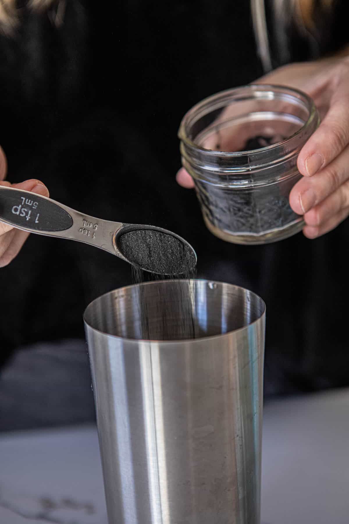 Woman adding activated charcoal to a cocktail shaker.