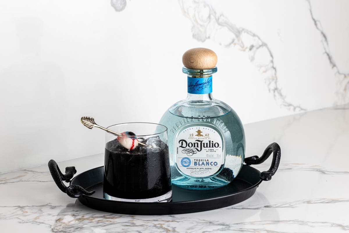 A black Halloween tray with a black cocktail drink next to a bottle of Don Julio tequila. 