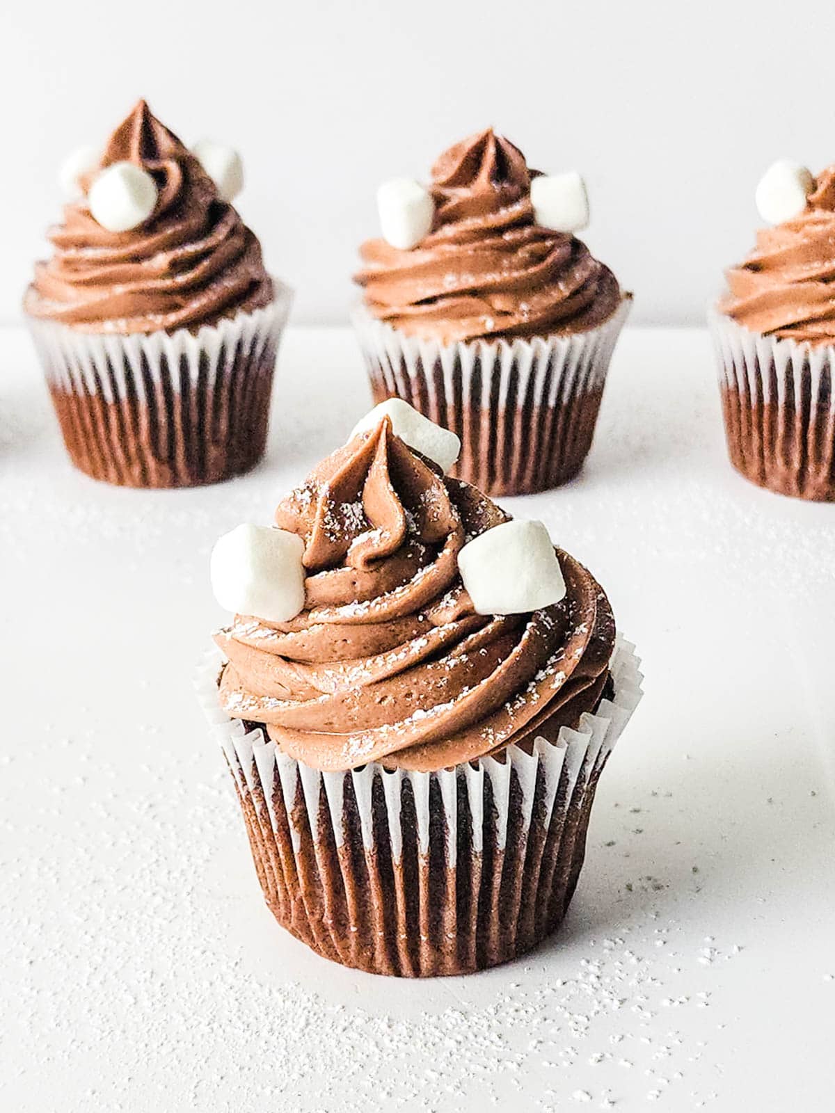 Close up of a hot cocoa cupcake topped with hot chocolate frosting powdered sugar and mini marshmallows.